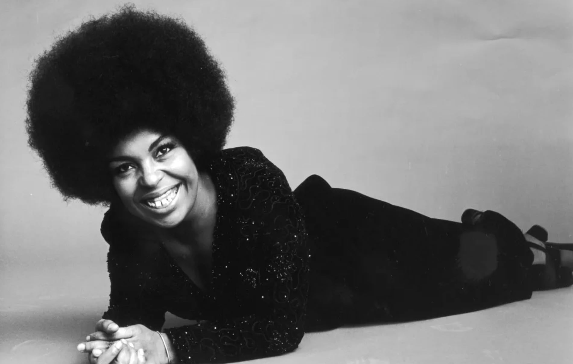 A woman with a large afro hairstyle smiles while lying on her stomach, wearing a long-sleeved, sparkly black dress and heeled shoes against a plain background.