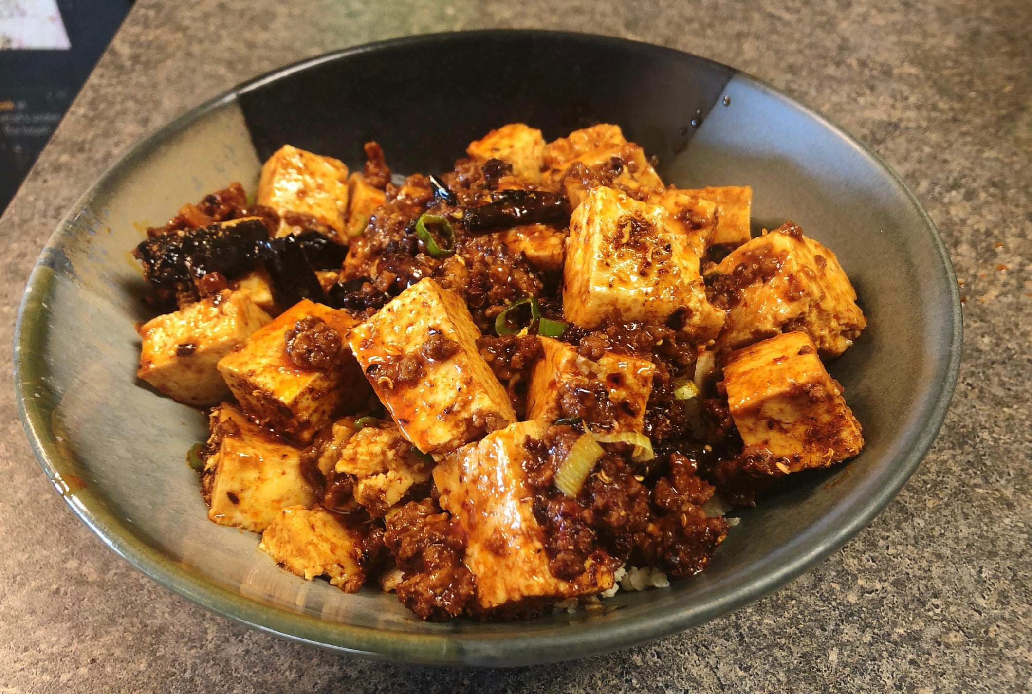 A bowl of mapo tofu featuring tofu cubes, ground meat, and spicy sauce with chopped green onions, served on a dark countertop.