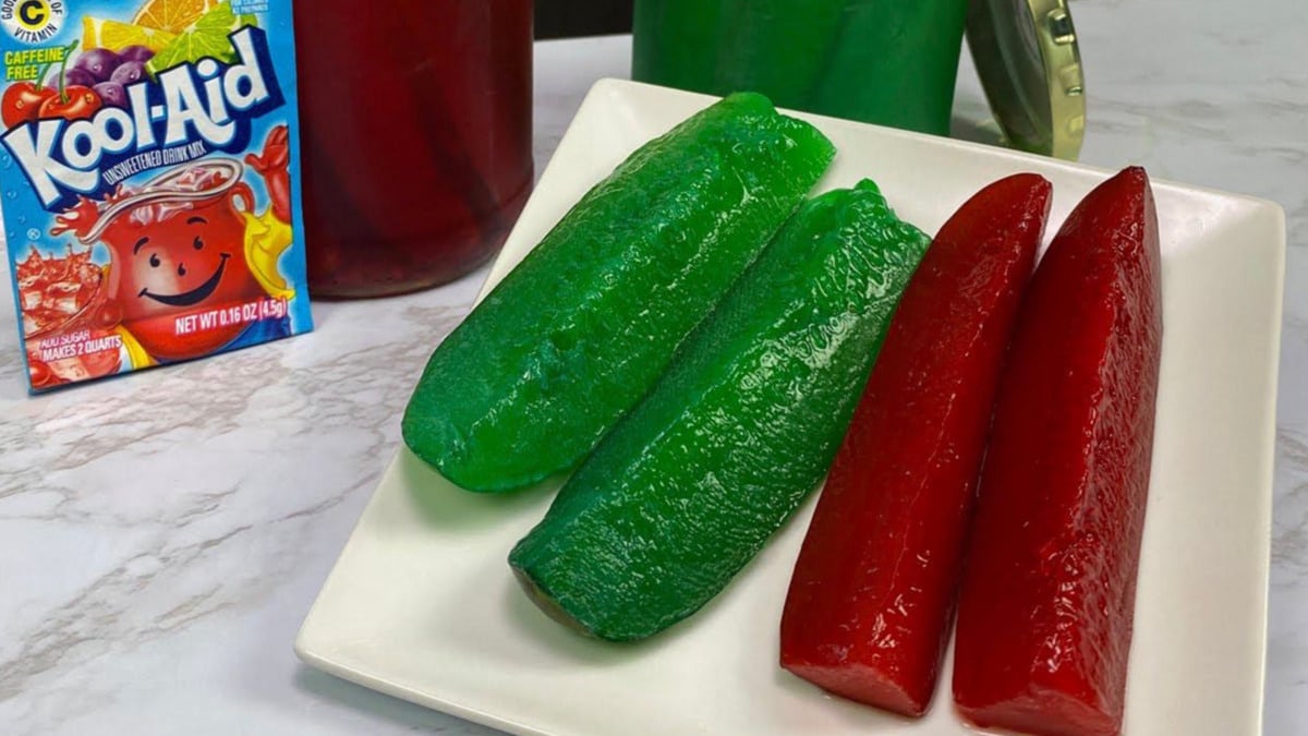 Four slices of pickles, two green and two red, are arranged on a white plate. A packet of Kool-Aid and jars of colorful drinks are visible in the background.
