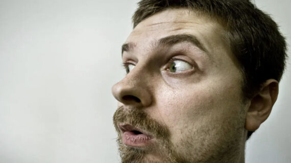 A man with light skin, brown hair, and a beard is looking to his left with wide eyes and an open mouth, appearing surprised or shocked. The background is plain and light-colored.