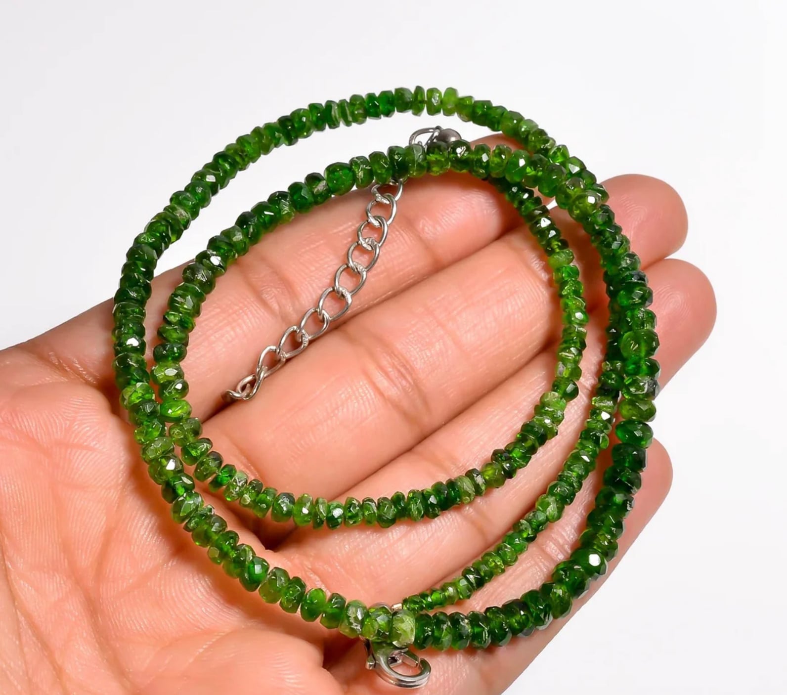 A hand holding a green beaded necklace with faceted stones and a silver chain extender.