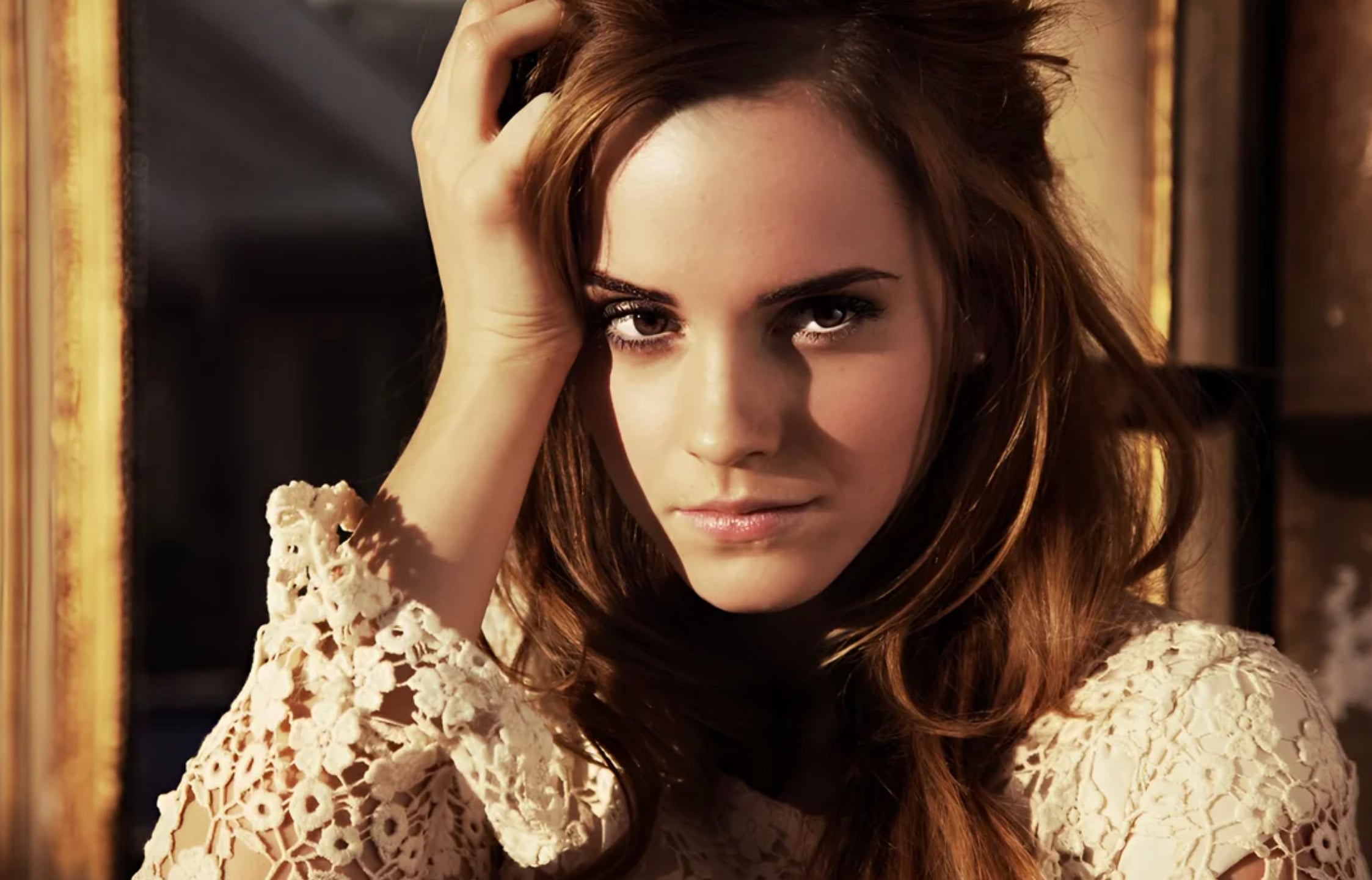 A woman with long, wavy brown hair gazes intensely at the camera. She wears a cream-colored, lace-patterned top and rests one hand on her head, with soft lighting highlighting her face.