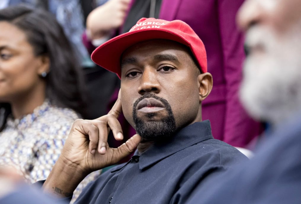 A man with a beard wearing a red "Make America Great Again" cap and a black shirt sits among other people, resting his face on his hand and looking directly at the camera.