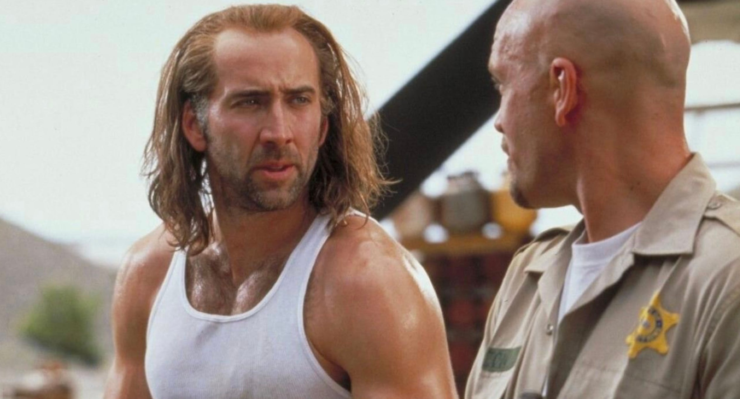 Two men stand facing each other; one with long hair and a white tank top, the other in a beige sheriff’s uniform. They appear to be having a serious conversation outdoors.