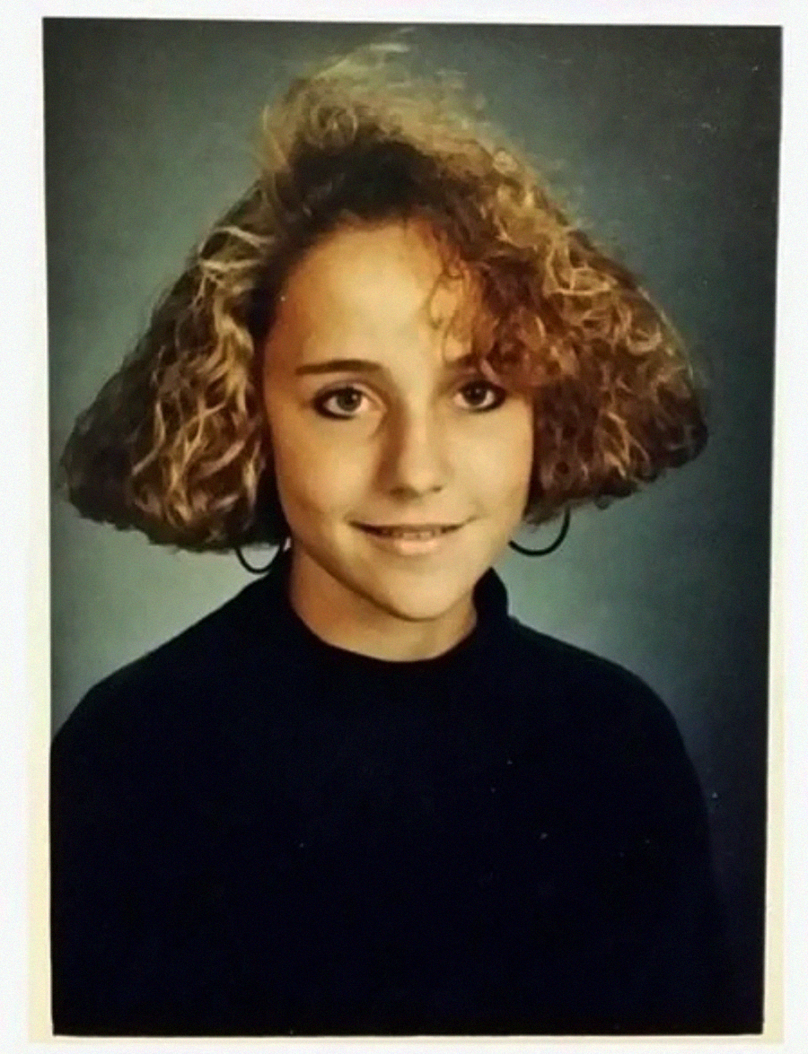 A young woman with curly, shoulder-length hair and hoop earrings smiles slightly in a studio portrait with a blue-gray background, wearing a dark top.