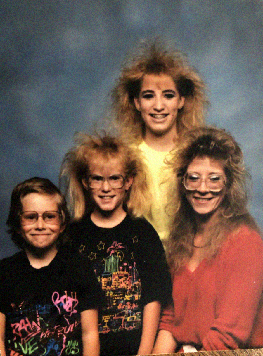 Four people with big, teased 1980s hairstyles and large glasses pose for a studio portrait against a blue background. They wear colorful and casual clothing, and all are smiling at the camera.