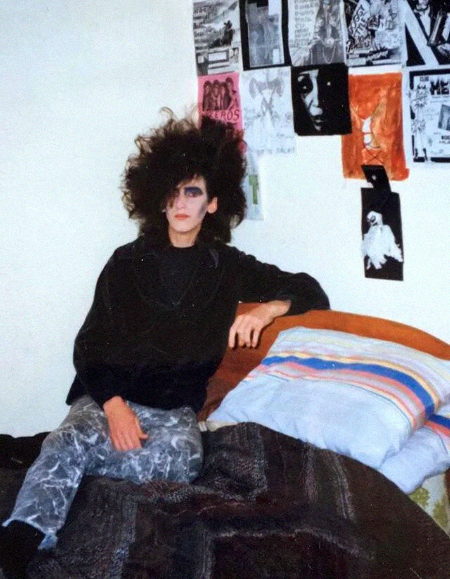 A person with teased, voluminous black hair and dramatic makeup sits on a bed, leaning against a wall decorated with posters and black-and-white prints. The bedding is striped and dark-colored.