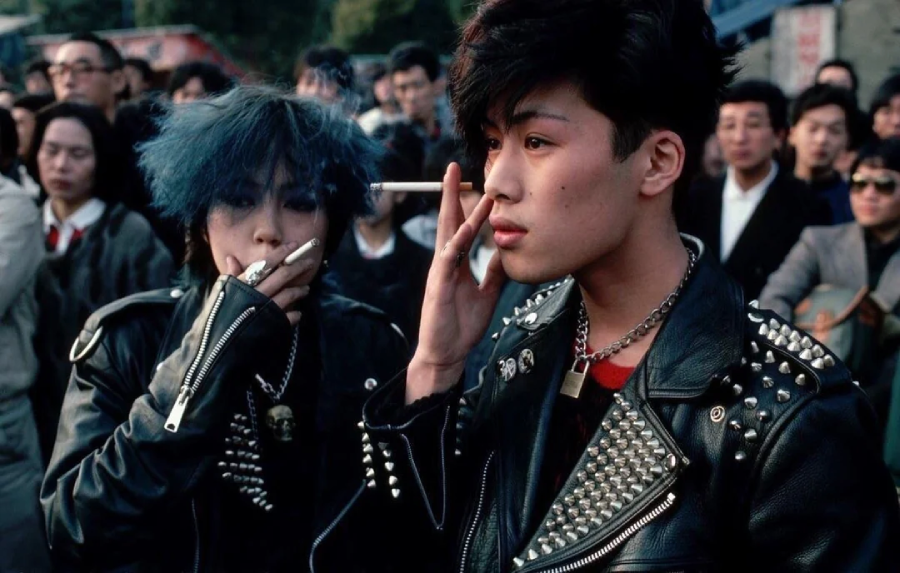 Two young people in studded black leather jackets stand in a crowd. One has spiky blue hair and holds a cigarette; the other touches their cheek, both with punk style. The background shows a mix of people watching them.