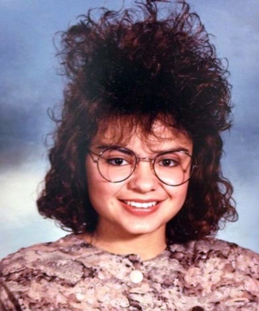 A young woman smiles in a school portrait, wearing large round glasses and a patterned top. Her curly dark hair is styled high with lots of volume, reflecting a vintage 1980s or early 1990s fashion.