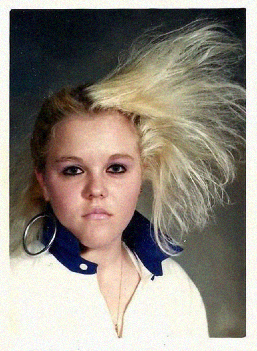 A young woman with bleached blonde hair styled dramatically to one side, wearing a white collared shirt with blue accents, large hoop earrings, and dark eye makeup, poses for a portrait against a neutral background.