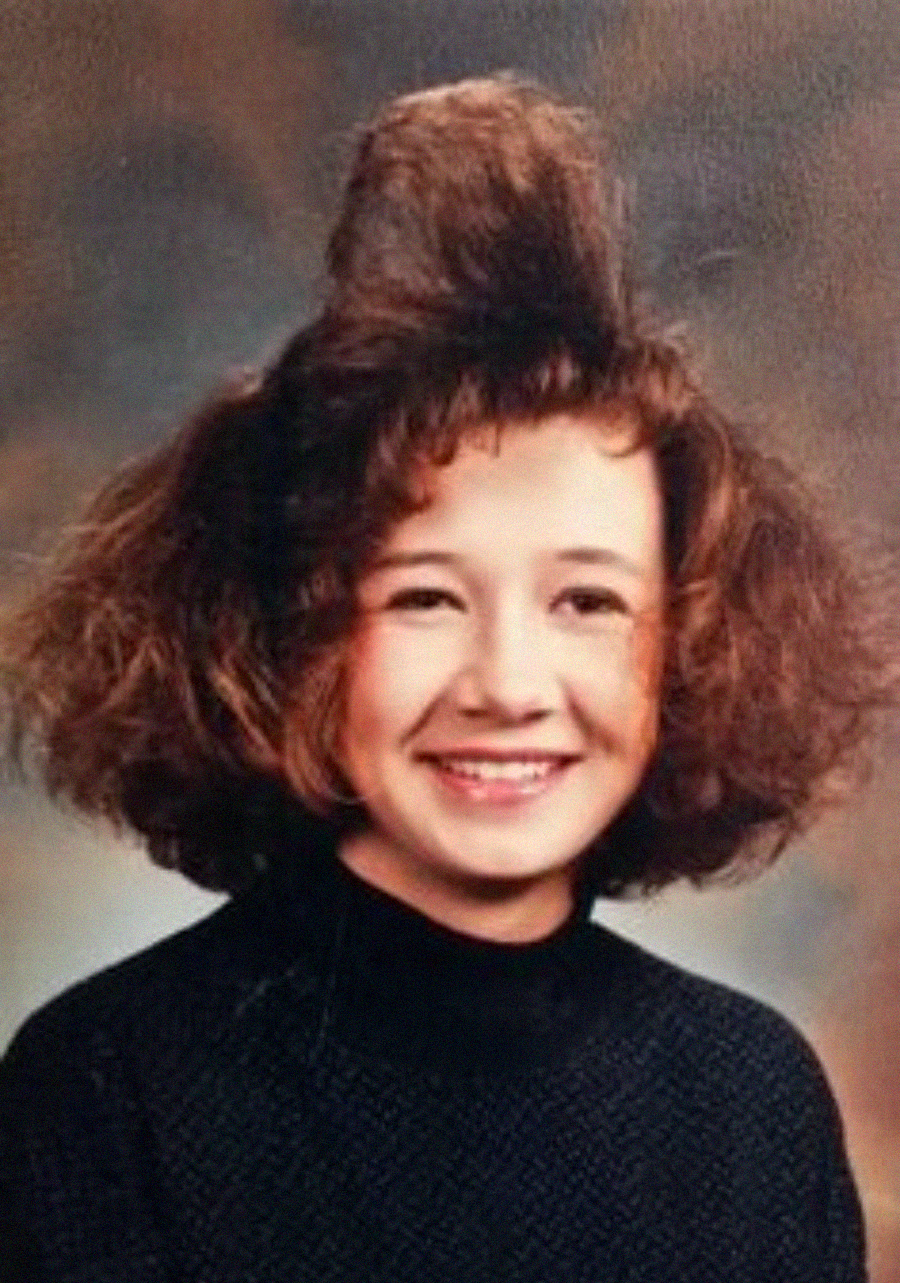 A young girl smiles in a school portrait, wearing a black sweater. Her brown hair is styled in a voluminous, teased fashion with a tall section sticking straight up on top. The background is softly blurred.