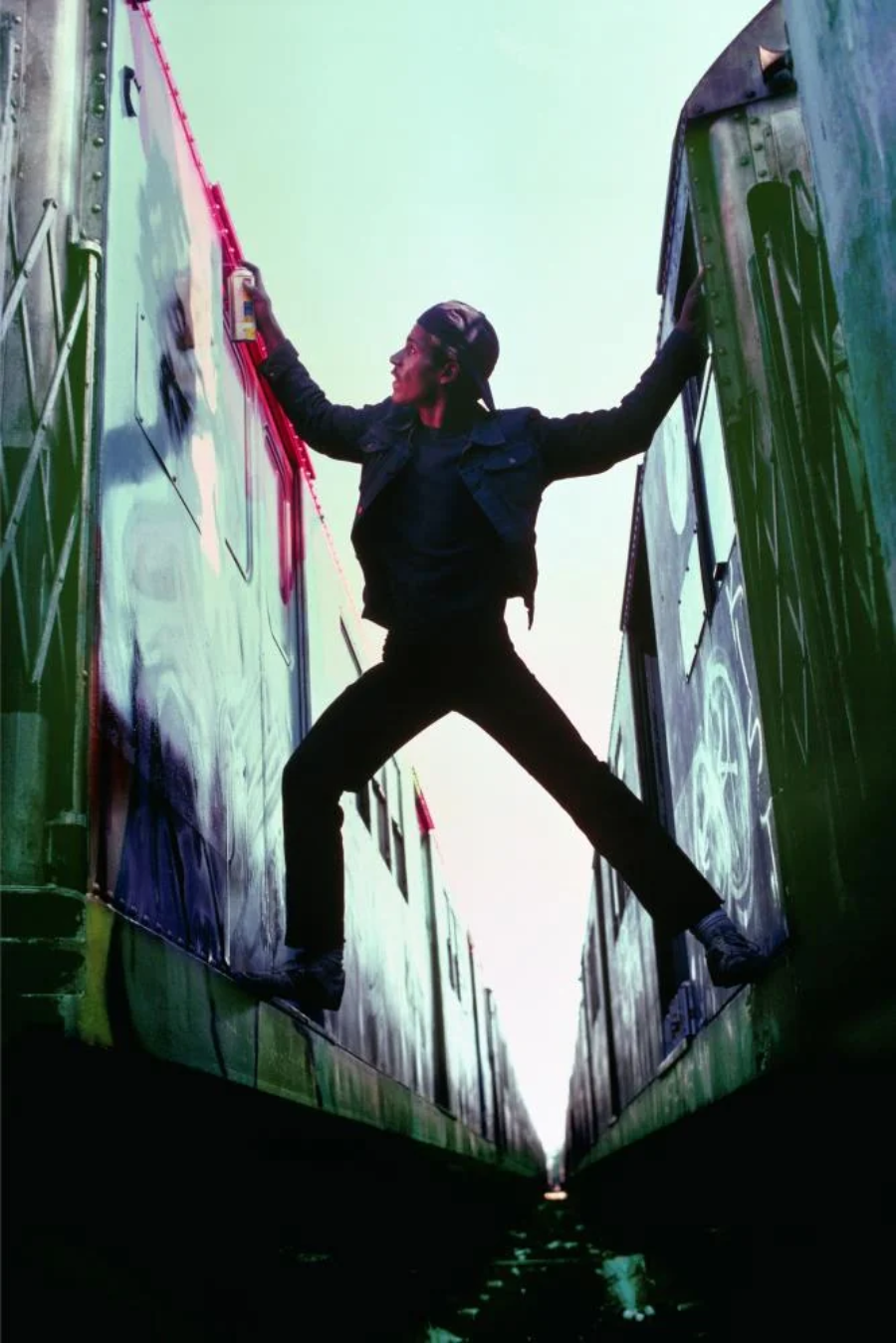 A person in dark clothing and a cap spray-paints graffiti on the side of a train while standing between two trains, balancing with one foot on each train car. The perspective is from below, emphasizing the action.
