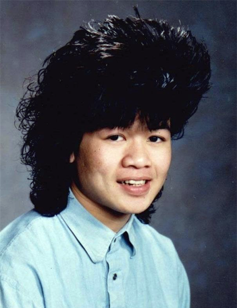 A young person with a large, voluminous, curly mullet hairstyle and wearing a light blue collared shirt poses for a studio portrait against a gray background.