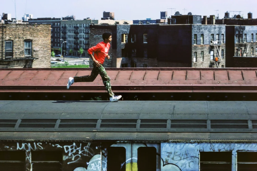 A person in a red jacket and camouflage pants runs on top of a graffiti-covered subway train, with brick apartment buildings and a cityscape in the background.