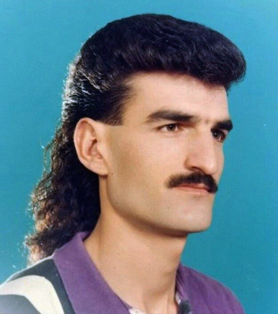 A man with a mullet hairstyle and a mustache poses for a studio portrait against a blue background, wearing a purple collared shirt with white and black stripes.