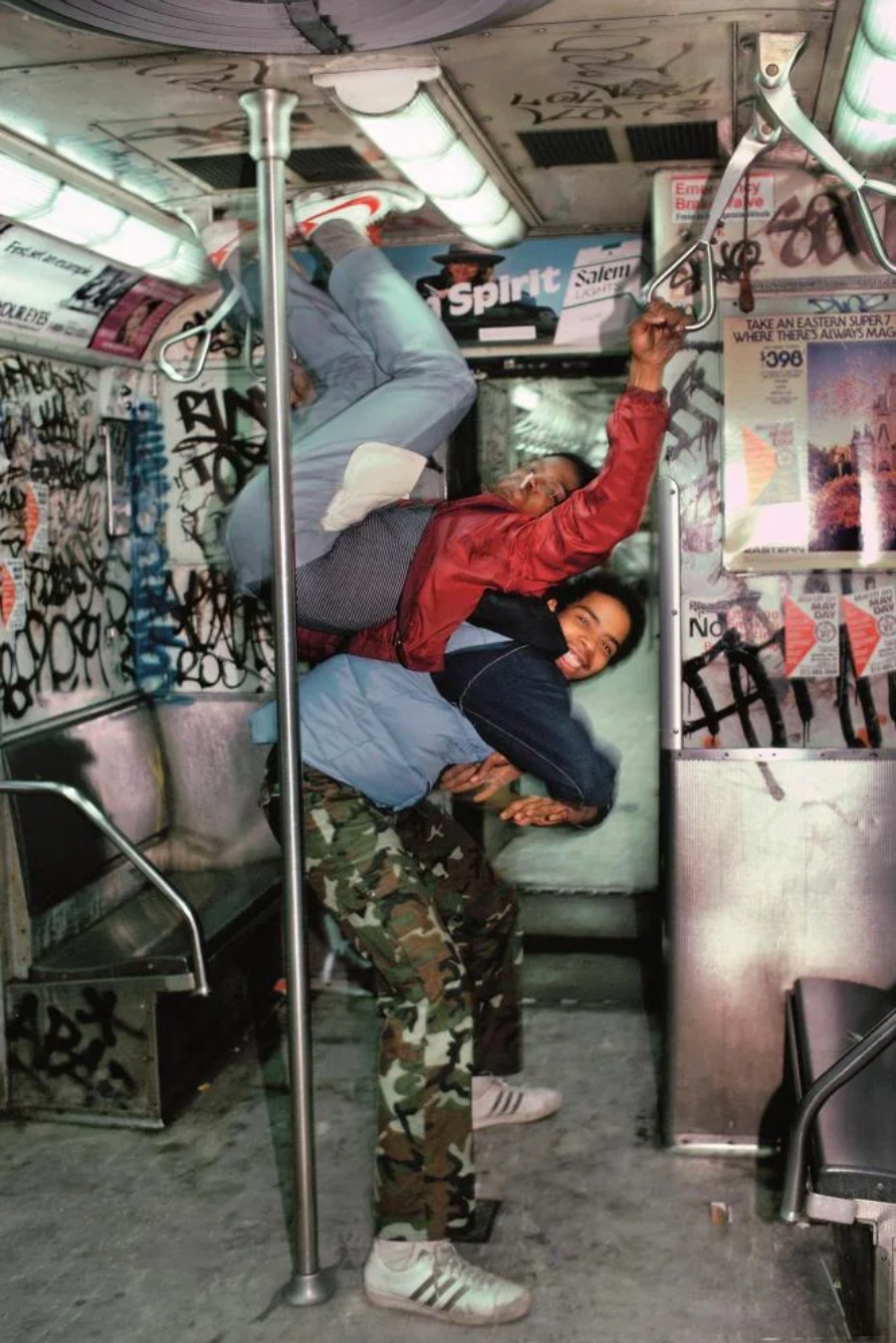 Two young men pose playfully in a graffiti-covered subway car; one hangs upside down from a pole while the other supports him, both smiling and wearing casual streetwear and sneakers.