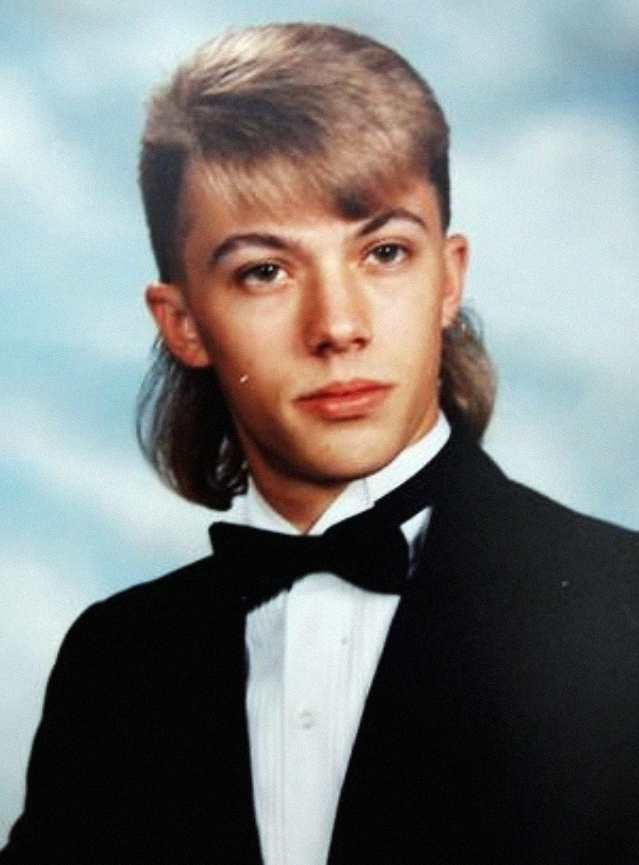 Young man with a mullet hairstyle wearing a black tuxedo jacket, white shirt, and black bow tie, posing for a formal studio portrait against a blurred blue and white background.