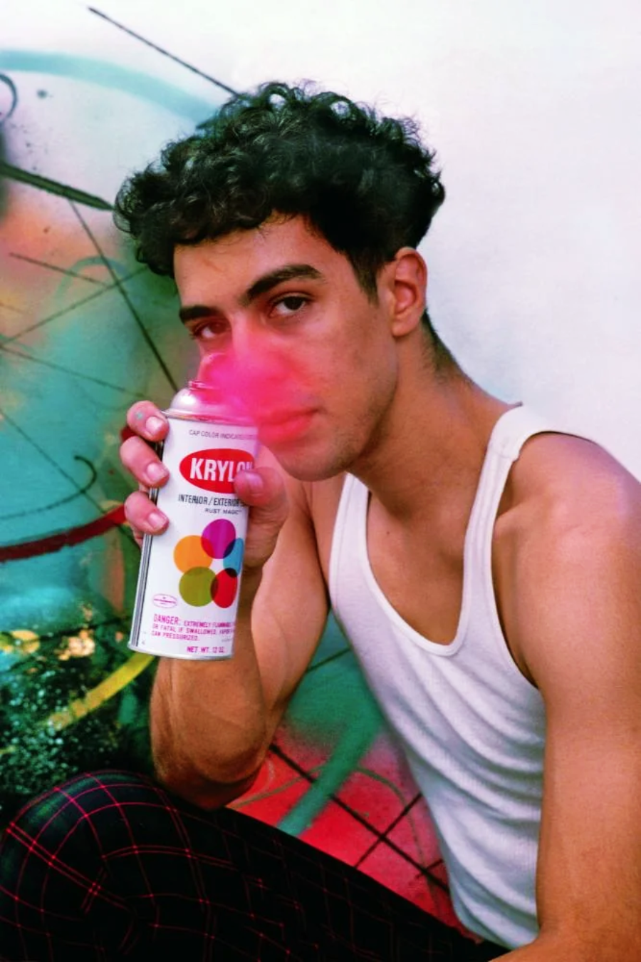 A young man in a white tank top holds a can of Krylon spray paint, spraying pink paint near his face. He sits in front of a colorful, graffiti-covered wall.