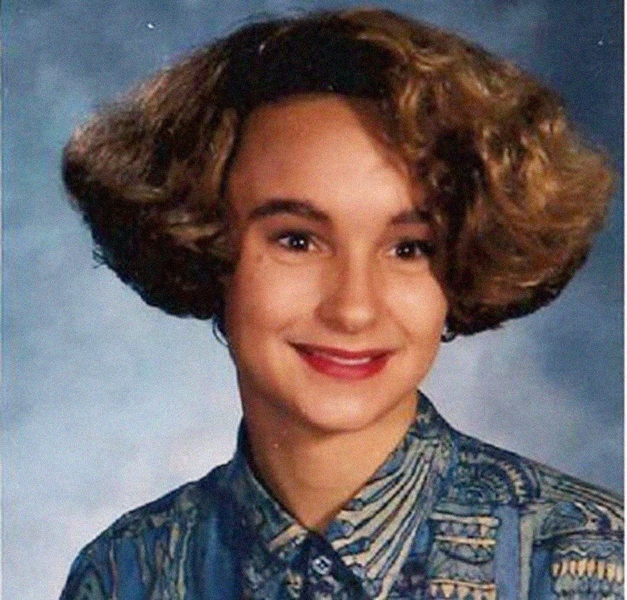 A young person with short, curly brown hair smiles at the camera in a school portrait. They wear a patterned button-up shirt and have a neutral background behind them.
