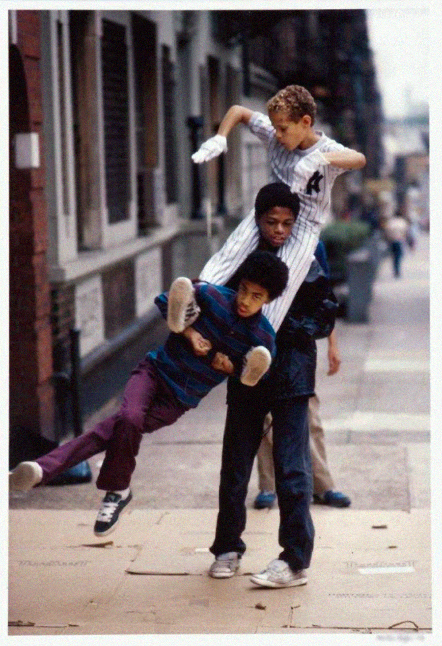 Three boys play on a sidewalk; one boy is sitting on another’s shoulders while a third boy swings by holding onto them. Another child walks behind them on the city street. The scene is lively and playful.