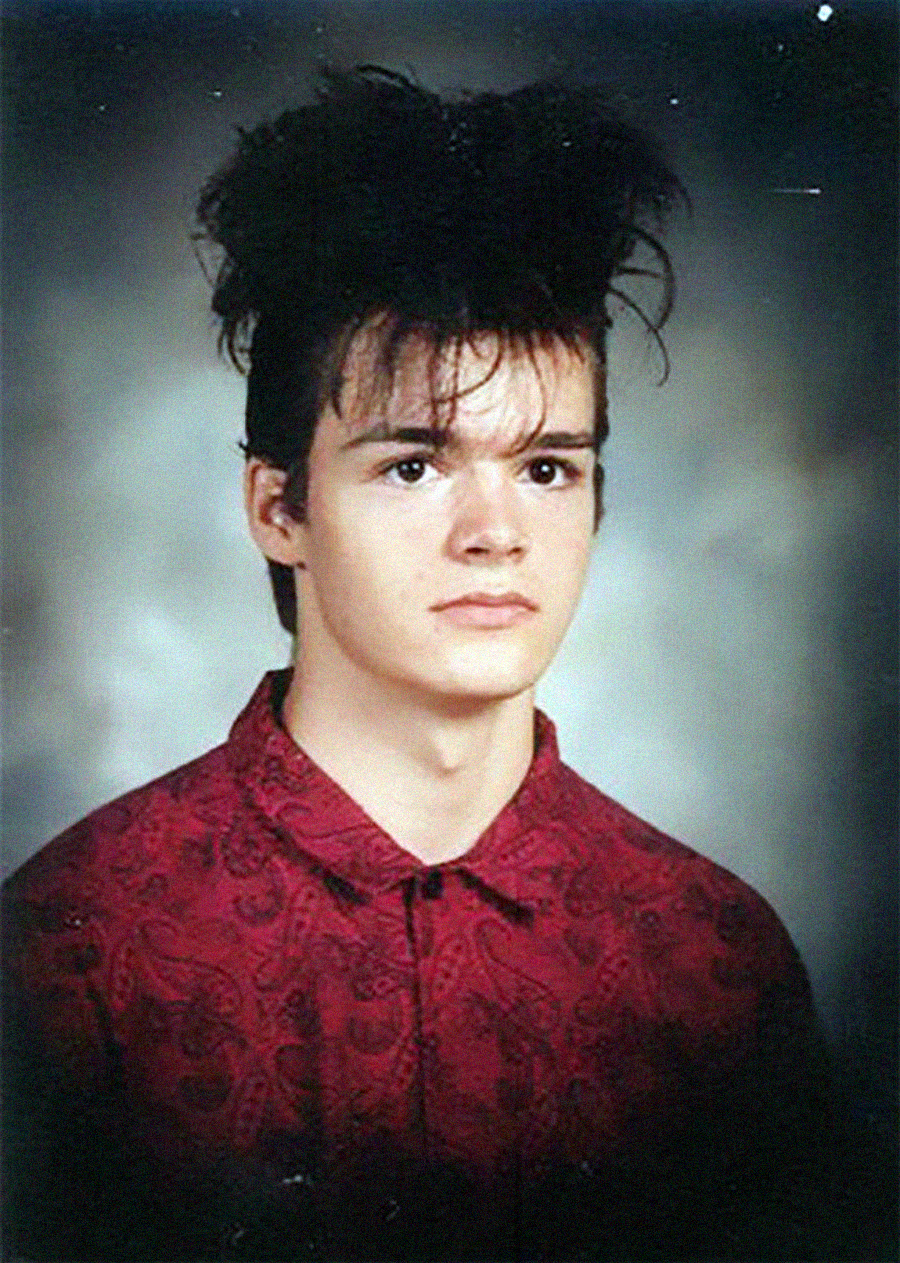 A young person with pale skin and tousled, voluminous dark hair wears a red patterned shirt and looks forward with a neutral expression against a gray studio background.