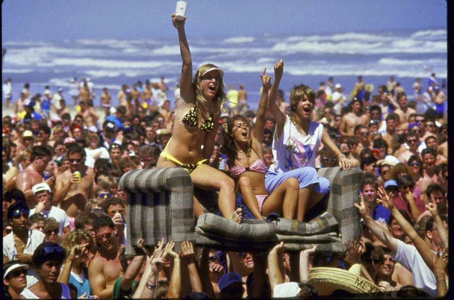 Three women sit atop an old couch being carried by a crowd at a packed beach party. One woman in a bikini holds up a drink and cheers, while the beach and ocean are visible in the background.