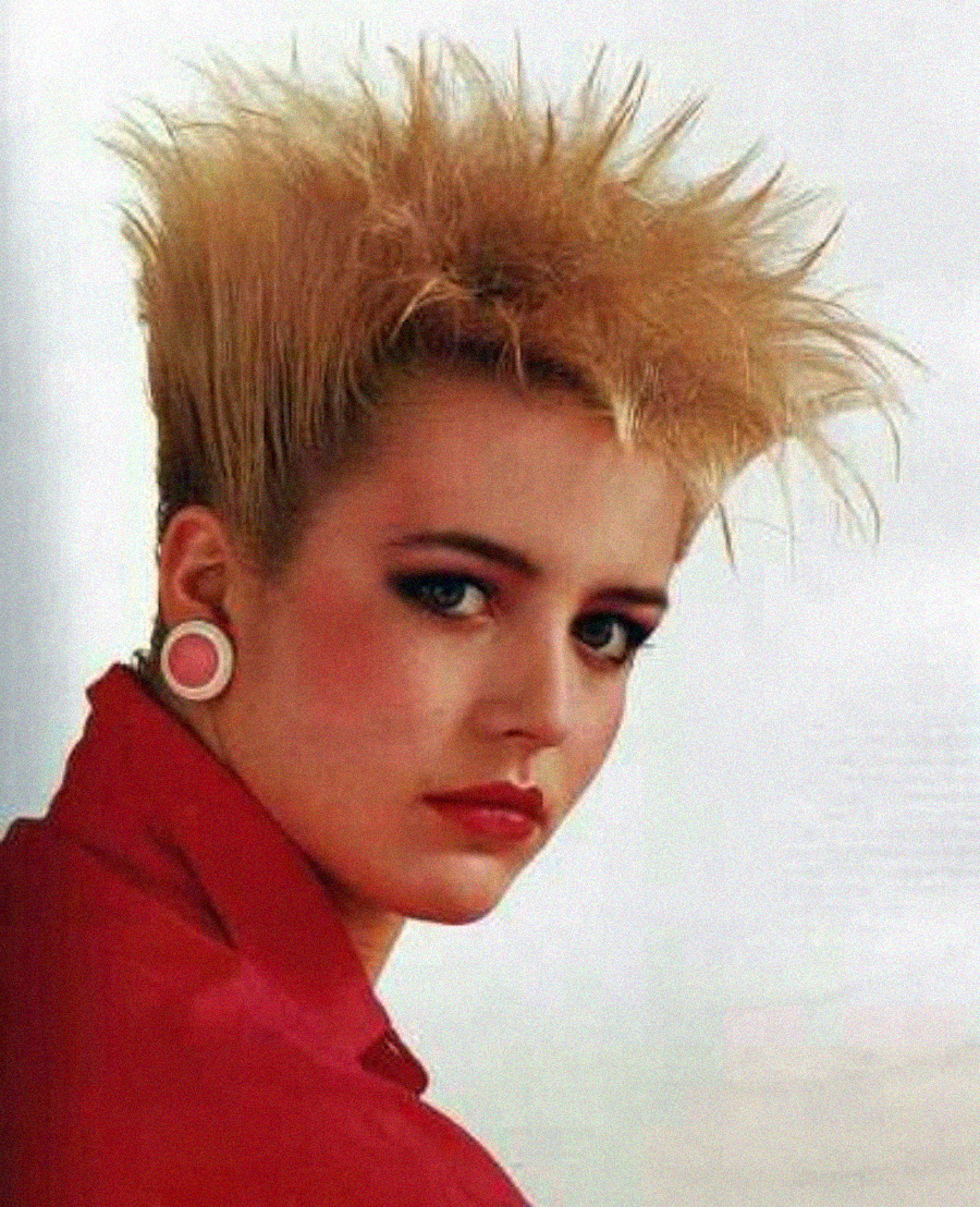 A woman with spiky, bleached blonde hair and large round earrings gazes at the camera. She is wearing a red top, red lipstick, and has a neutral expression against a light background.