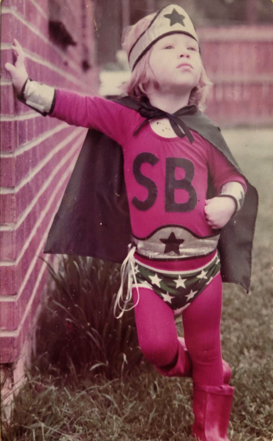 A young child poses confidently outdoors in a handmade superhero costume with a cape, boots, headband, and an outfit featuring stars and the letters "SB" on the shirt, leaning against a brick wall.