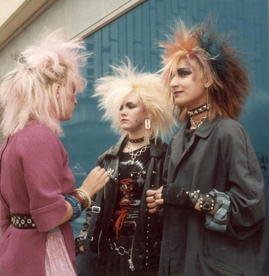 Three people with brightly colored, teased punk hairstyles and dramatic makeup stand talking on a city street, wearing leather jackets, spikes, chains, and bold accessories in front of a blue wall.