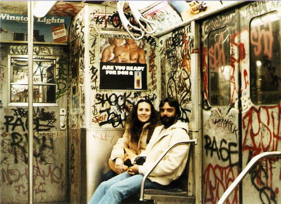 A man and woman sit together and smile in a graffiti-covered subway car. Colorful tags and spray paint cover the walls, doors, and ceiling. An old advertisement and a "Winston Lights" sign are visible above them.