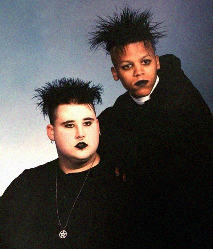 Two people with spiked black hair, dramatic black lipstick, and dark clothing pose together against a plain background. One wears a pentagram necklace; both have a goth style.