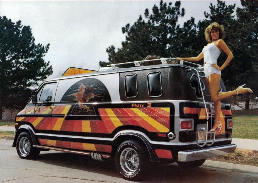 A woman in a white outfit poses on the rear ladder of a custom-painted van with colorful stripes and a fantasy mural, parked by trees on a cloudy day.