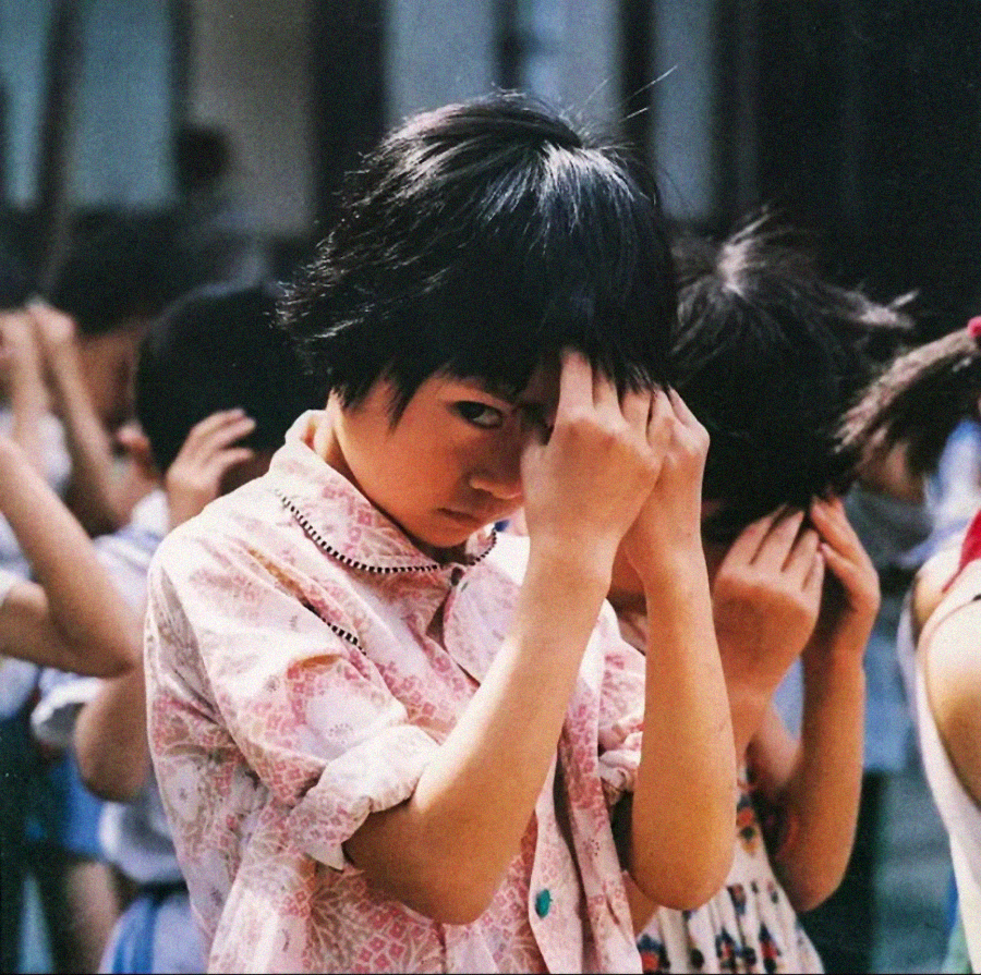 A young girl in a pink patterned shirt stands in a group, parting her dark hair and looking at the camera while others around her also check their hair, possibly during a lice inspection.