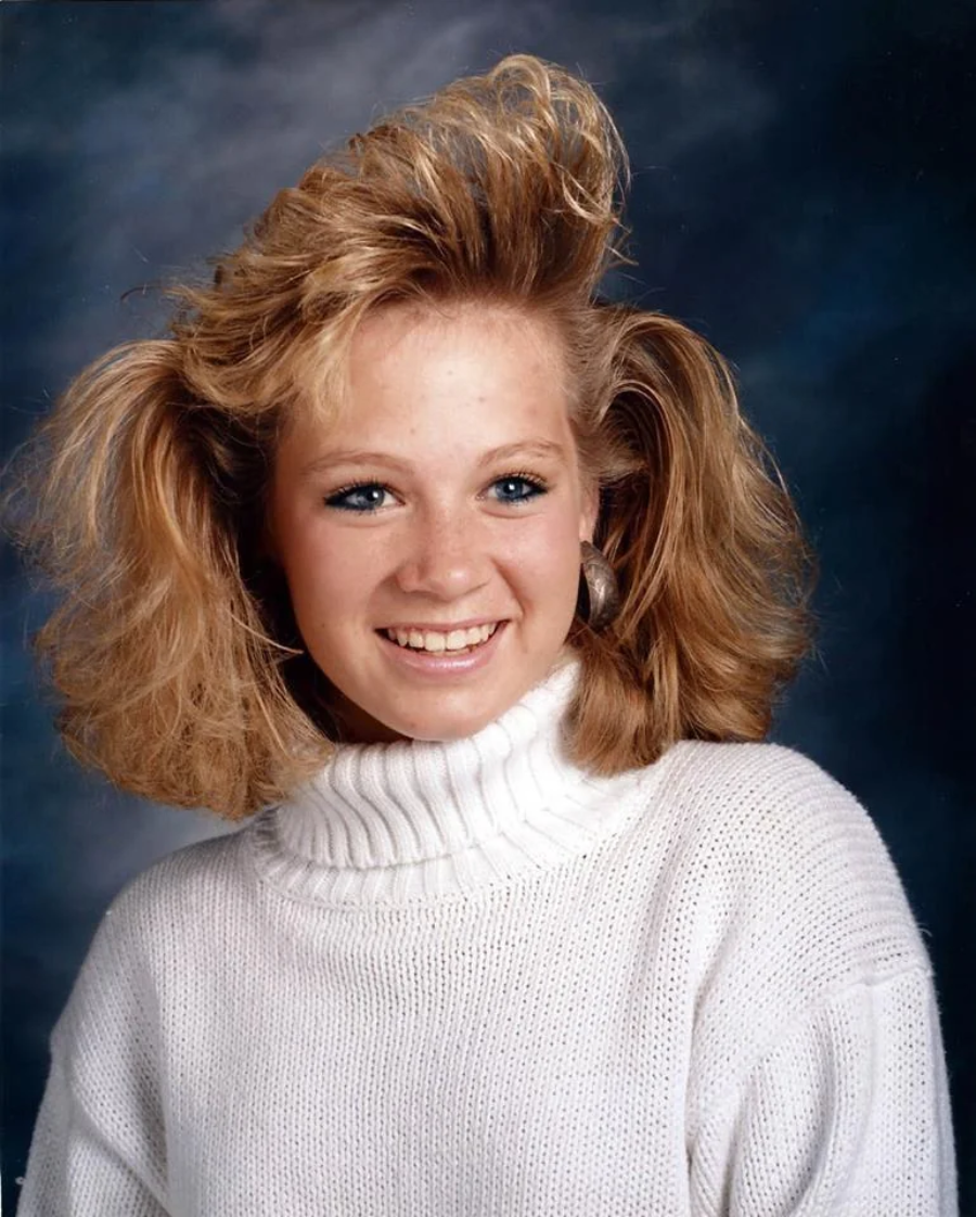 A teenage girl with blond, voluminous 1980s-style hair smiles at the camera. She wears a white turtleneck sweater and large earrings, with a blue and black studio backdrop behind her.