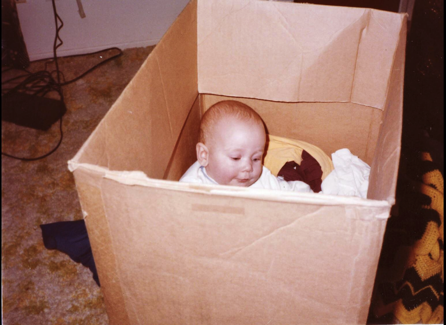 A baby sits inside a large cardboard box on a carpeted floor, surrounded by clothes. Electronic cords and a wall outlet are visible in the background.
