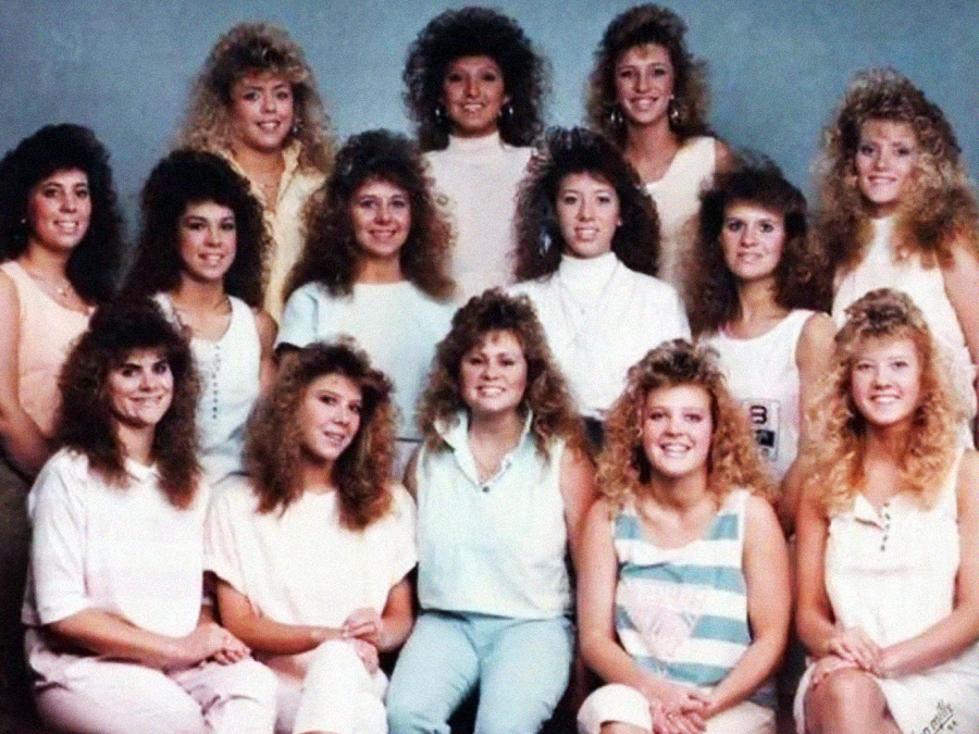 A group of thirteen women with big, voluminous 1980s-style hair pose together in pastel-colored outfits against a plain backdrop, smiling for a group photo.