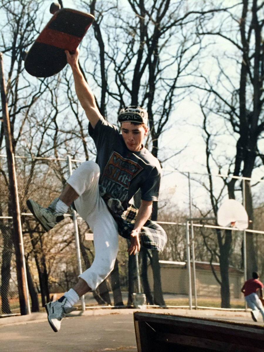 A skateboarder wearing a beanie and T-shirt grabs his board midair while jumping off a ramp in an outdoor skate park surrounded by leafless trees and a chain-link fence.