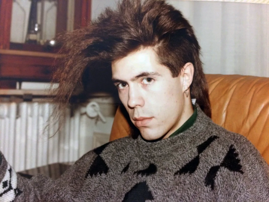 A young man with a mullet hairstyle and an earring sits on a tan leather chair, wearing a gray sweater with a black geometric pattern, looking seriously at the camera.