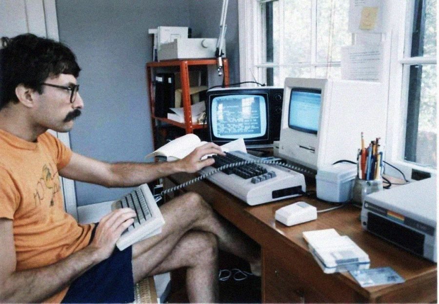 A man with glasses and a mustache uses a vintage computer setup with multiple monitors, a keyboard, and a printer on a cluttered desk in a bright, windowed room.