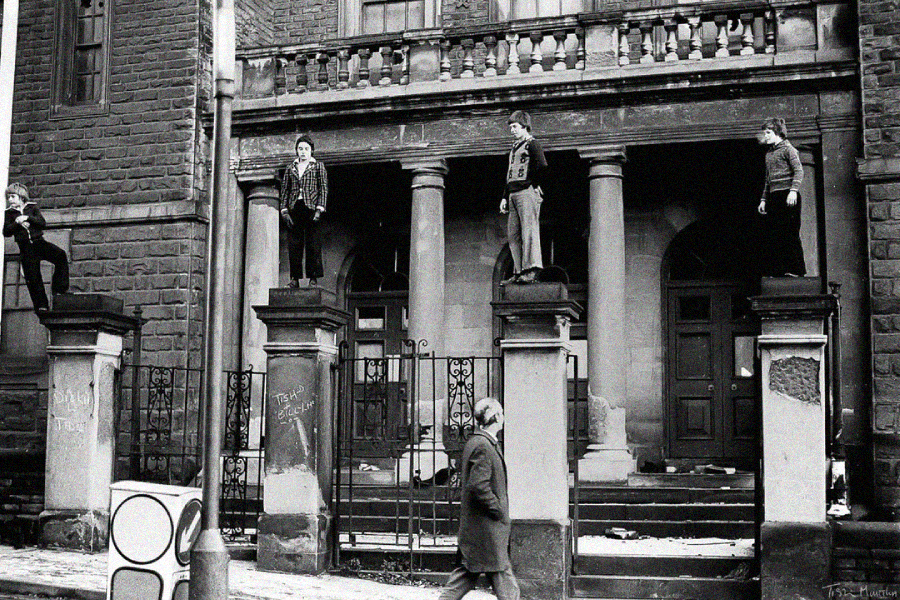 Four people stand atop stone pillars outside a worn, columned building, while a person walks by on the sidewalk below. The scene is in black and white, suggesting an old or vintage photo.