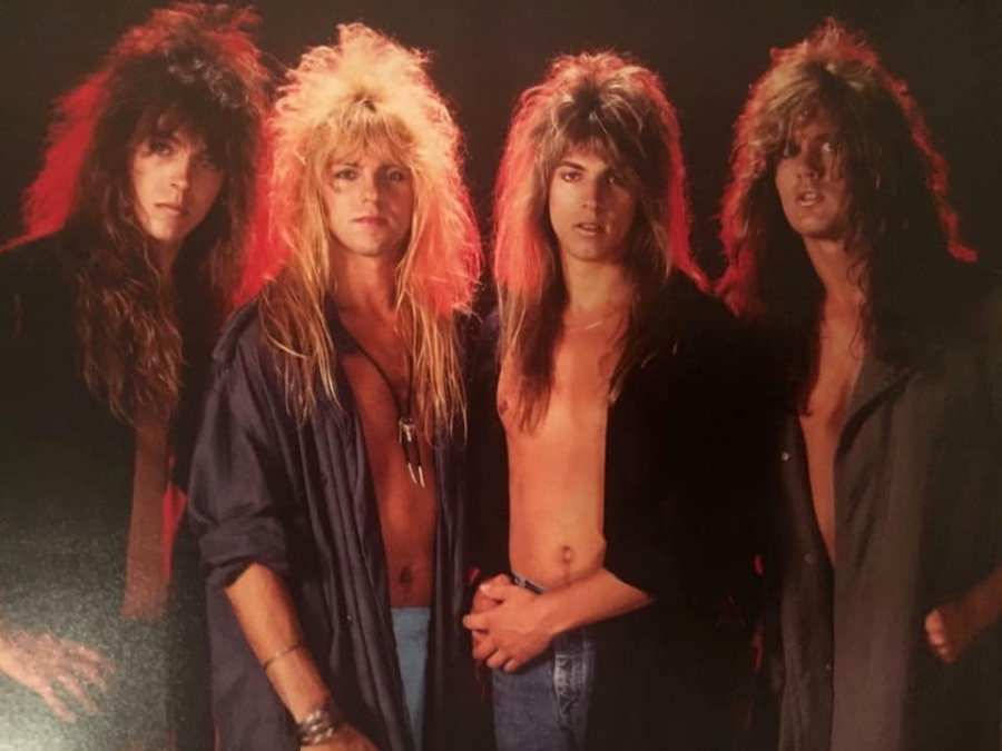 Four young men with long, teased 1980s-style hair pose together against a dark background with red lighting. They wear open shirts or jackets, exposing their chests, and have serious expressions.