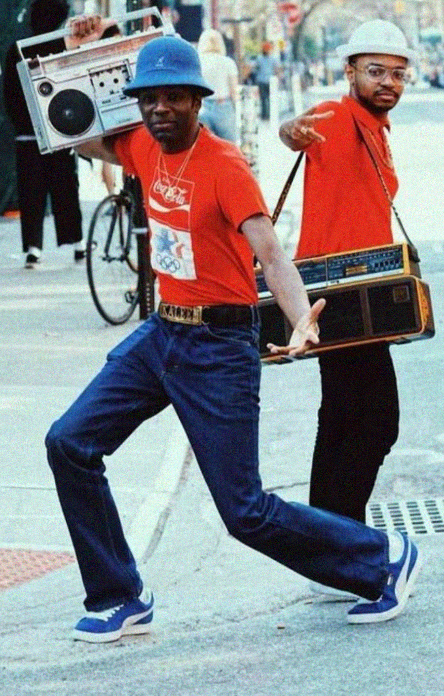 Two men in blue hats and red shirts strike dynamic poses on a city sidewalk, each carrying a large vintage boombox. One man steps forward energetically, while the other points toward the camera.