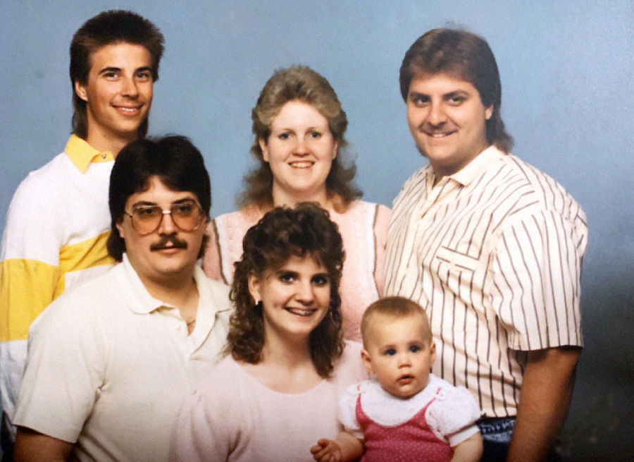 A group of five adults and one baby pose for a studio portrait, all wearing light-colored 1980s-style clothing and smiling at the camera. The adults have feathered hair and mullets. The baby is in a pink dress.