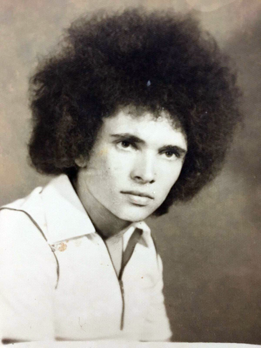 A young person with a large, curly afro hairstyle and a serious expression, wearing a light-colored collared shirt, poses for a vintage-style portrait against a plain background.
