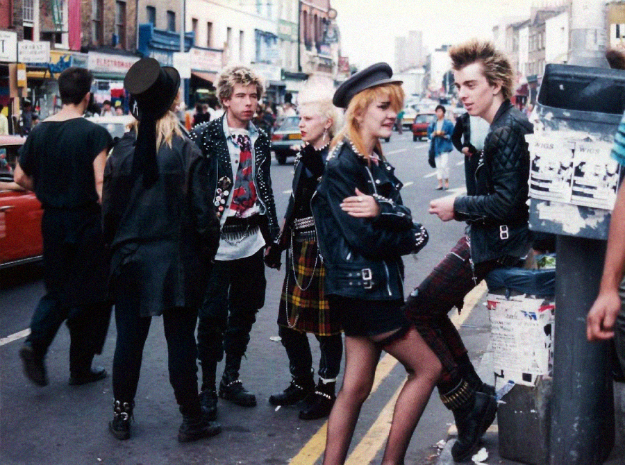 A group of young people in punk fashion—leather jackets, plaid skirts, spiked hair—stand and chat on a busy city street lined with shops and posters on a pole.
