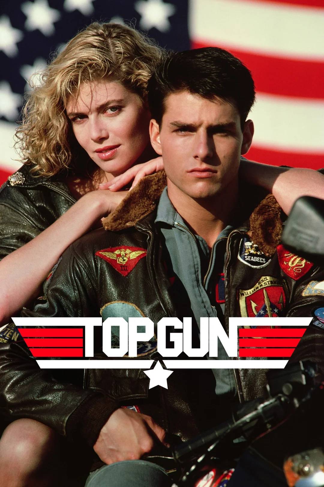 A man and woman in aviator jackets pose closely in front of a large American flag. The man is seated, looking serious, while the woman stands behind him, resting her arms on his shoulders. The "Top Gun" logo appears at the bottom.