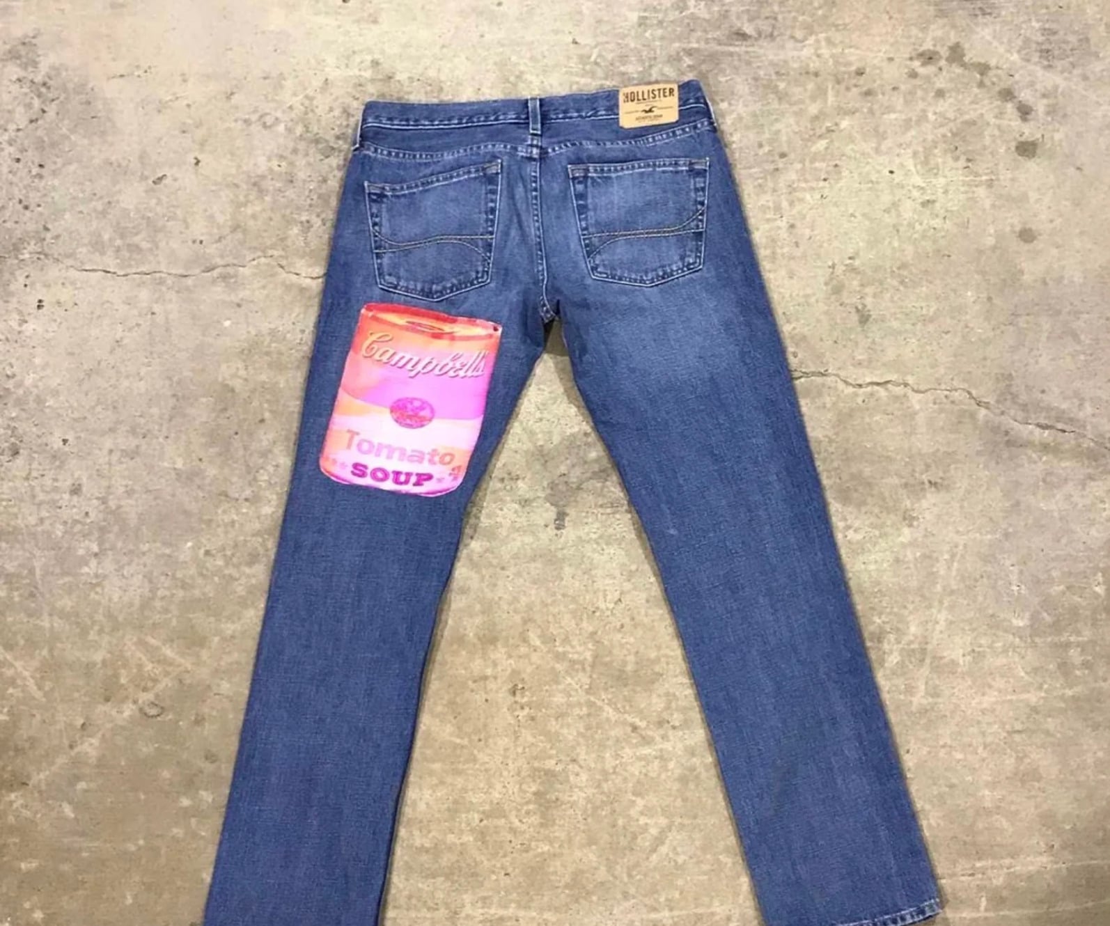 A pair of blue jeans lies flat on a concrete floor, featuring a large patch of a pink and orange Campbell’s Tomato Soup can on one back pocket.