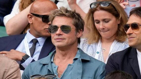 A man wearing sunglasses and a blue collared shirt sits among a crowd at an outdoor event. Other people near him are also wearing sunglasses and appear engaged in the event.