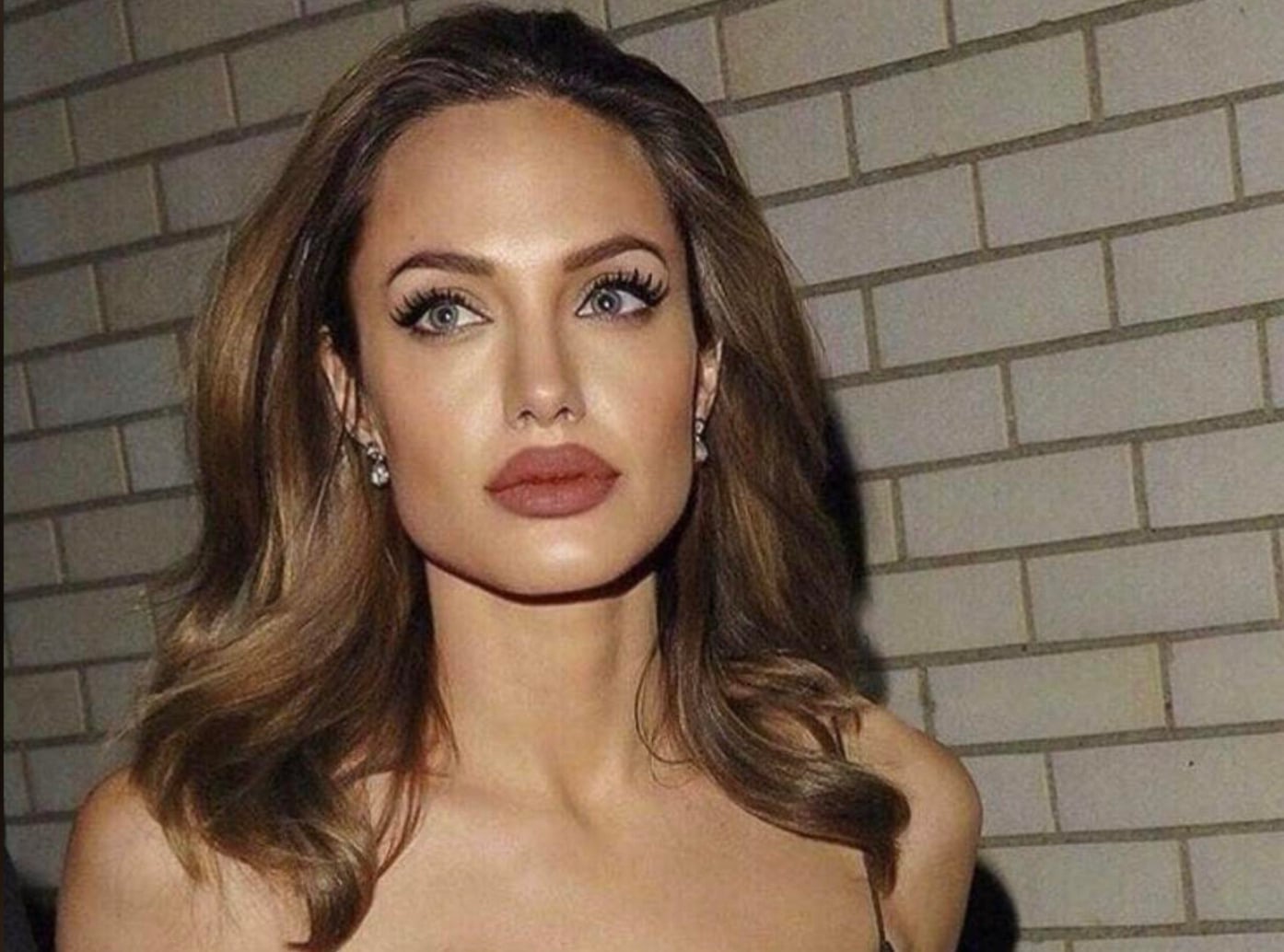 A woman with wavy brown hair, striking blue eyes, and bold red lipstick stands in front of a light brick wall, wearing earrings and a strapless top.