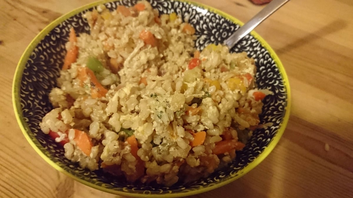 A patterned bowl filled with fried rice mixed with chopped vegetables, including carrots and bell peppers, sits on a wooden table with a metal spoon inside.